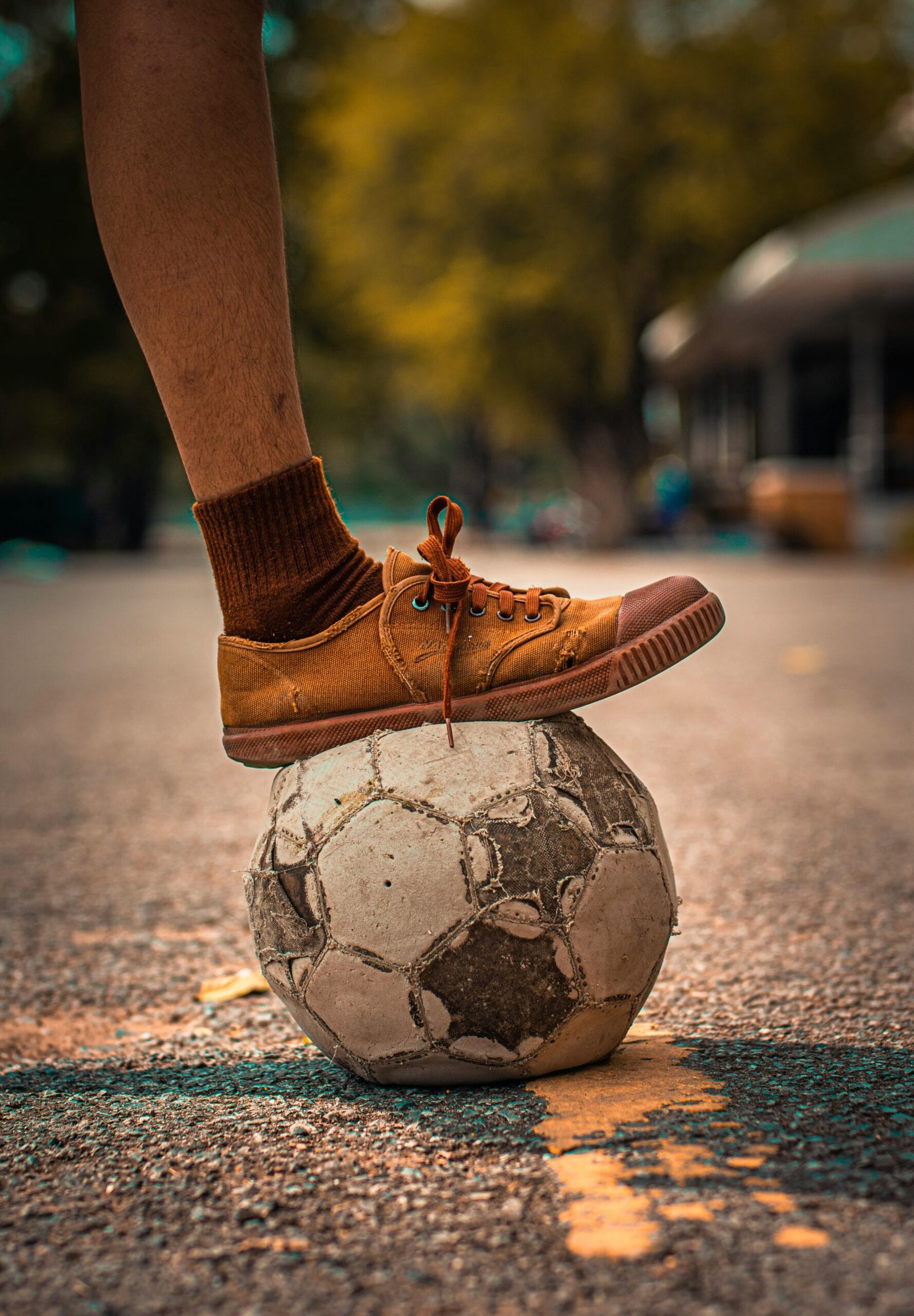 An aged soccer ball under a foot in casual footwear on an outdoor surface.